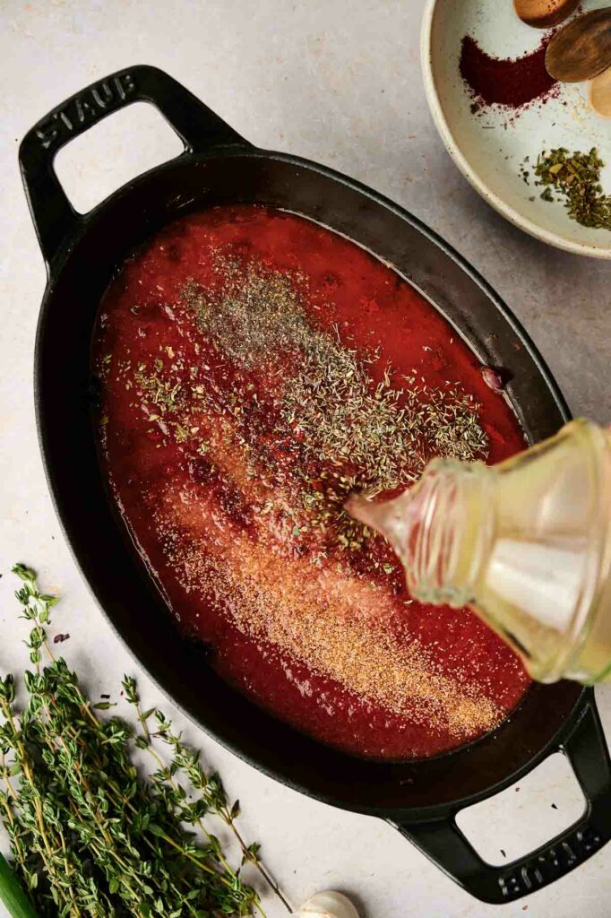 A ratatouille recipe pan with a sauce and herbs on it, and a bottle of broth being poured on it.