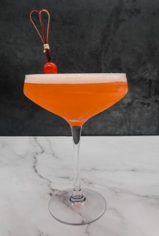 A homemade bright orange Aperol sour cocktail with a frothy top, with a cocktail stick with a maraschino cherry on.