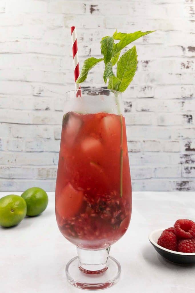 A freshly made raspberry mojito cocktail drink in a cocktail glass, with fresh mint garnish, a striped red straw, raspberries on a plate and two limes in the background.