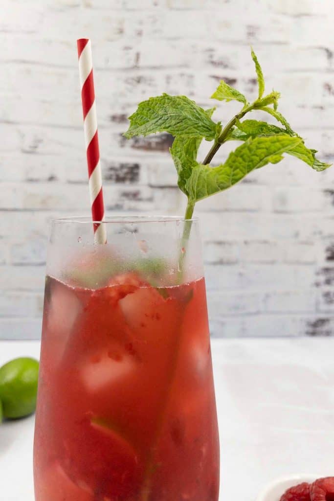 Close up shot of a homemade raspberry mojito cocktail with fresh mint garnish and a straw, a fresh lime and raspberries in the background.