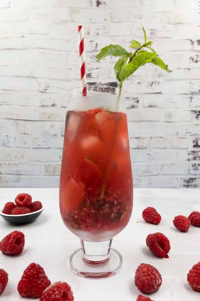 A cocktail glass of homemade raspberry mojito with a fresh mint garnish, striped red straw and fresh raspberries.