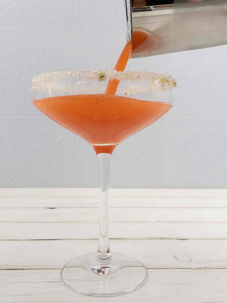 Pouring a homemade sweet strawberry daiquiri from a cocktail shaker into a coupe glass.