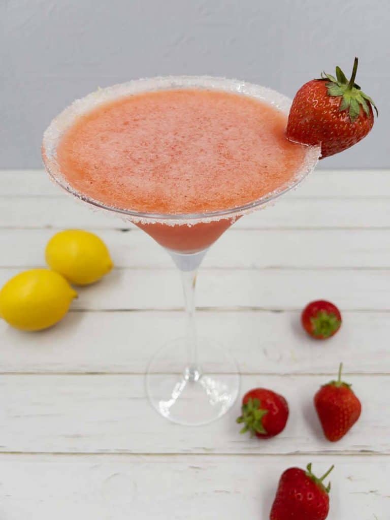 A refreshing glass of sweet strawberry martini with a sugared rim, strawberry garnish and strawberries and lemons surrounding it.