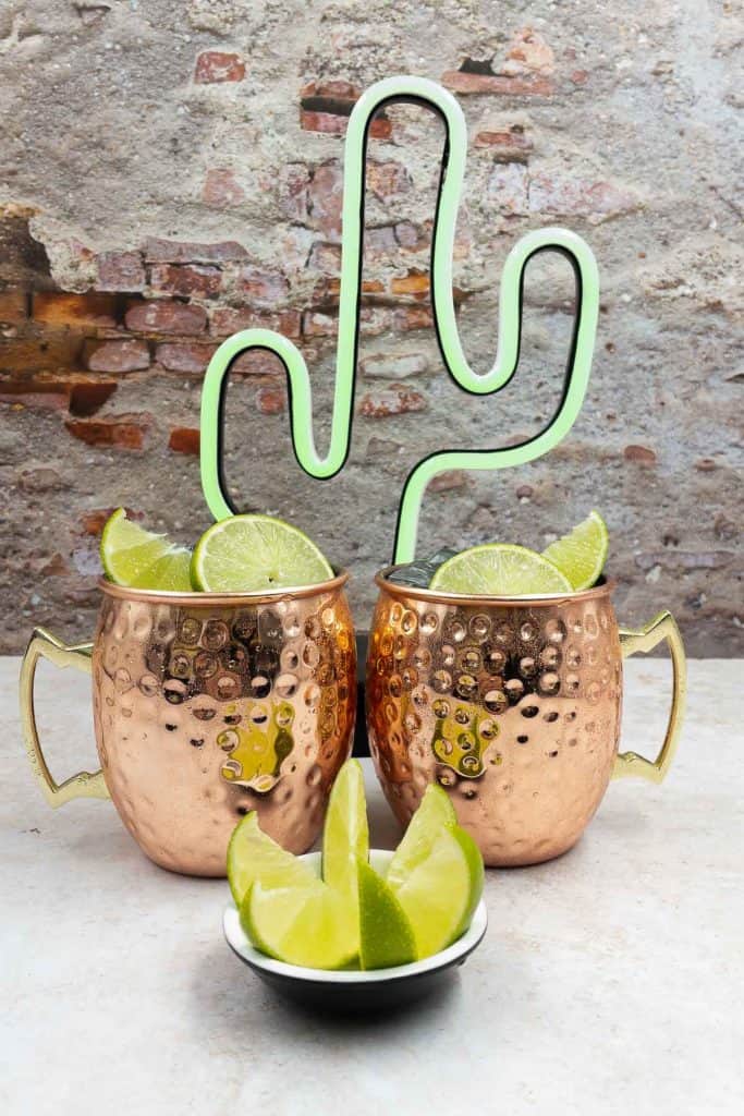 Two Mexican mules in copper mugs, with a lime wedge and a lime slice garnish, a neon cactus and a bowl of lime wedges.
