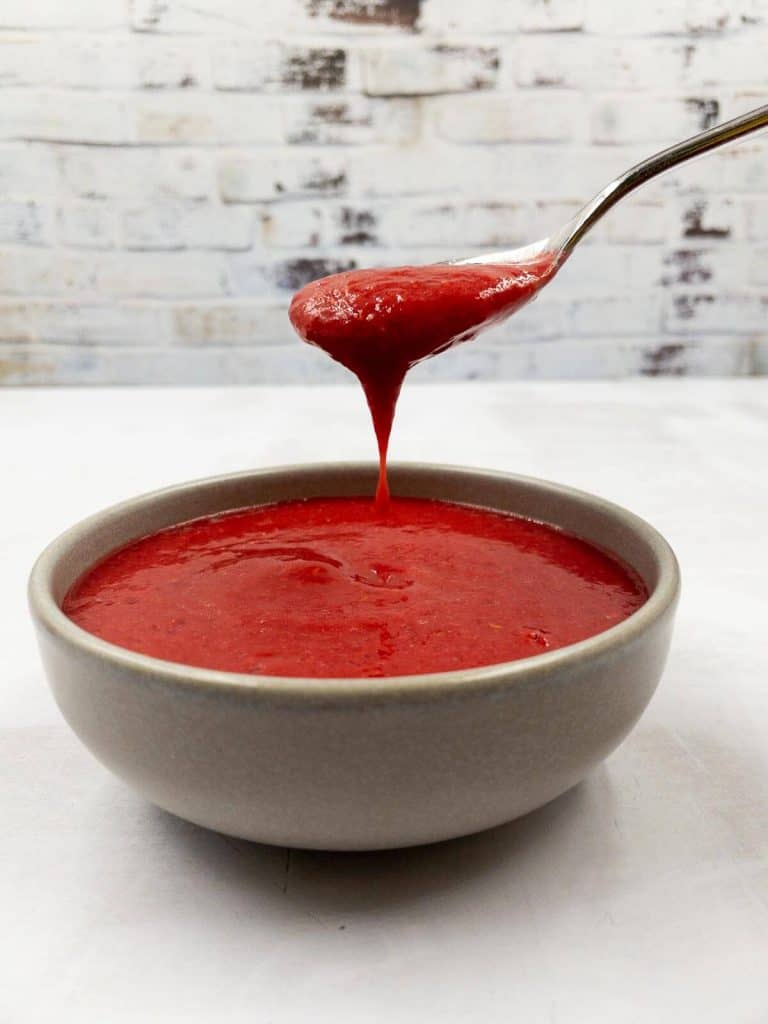 A bowl of fresh raspberry puree sauce and a spoon with puree dripping from it.