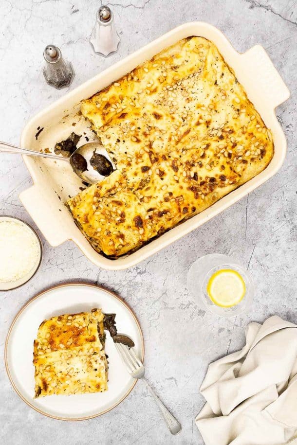 Overhead shot of delicious four cheese and mushroom lasagne al forno in a baking dish and a slice of the lasagna on a plate with a fork in it, salt and pepper pots, a bowl of parmesan and a glass of water with ice and a slice of lemon in it.