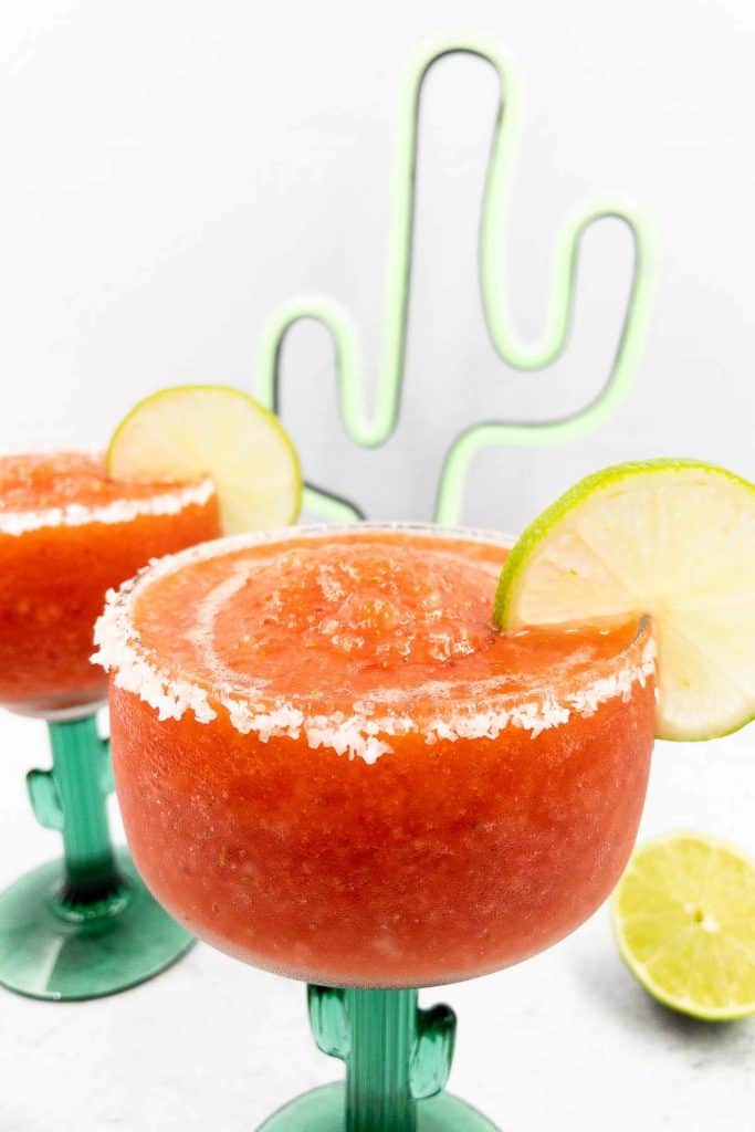 Two frozen strawberry margaritas ready to serve with lime slices and a cactus in the background.