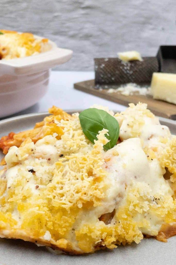 Pasta al forno baked and on a plate, in a baking dish and with grated parmesan cheese in the background.