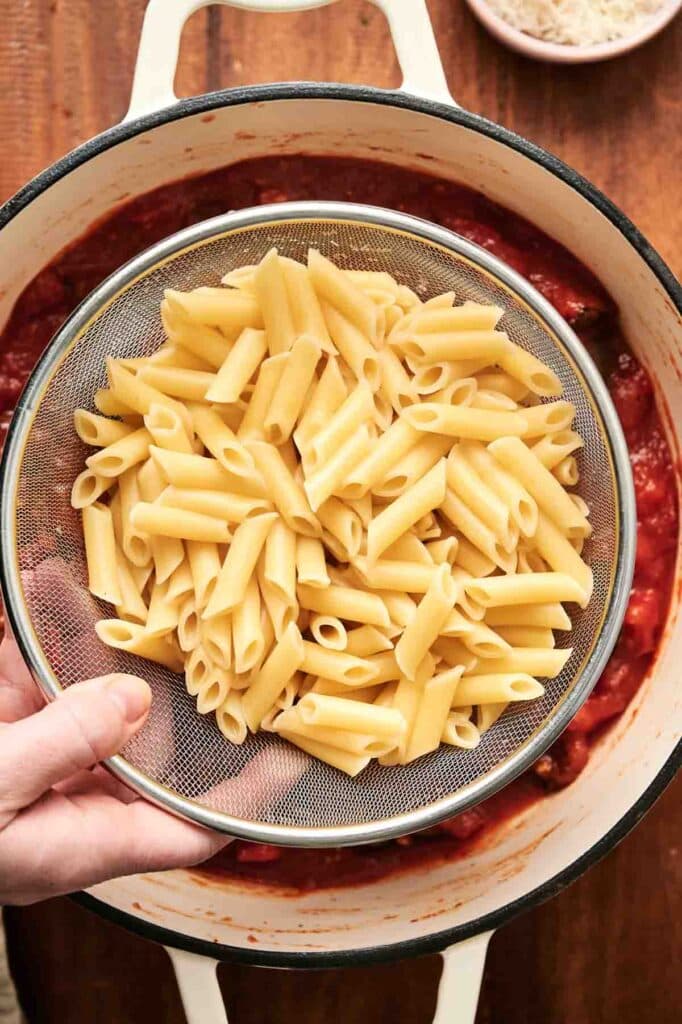 A hand holds a sieve with cooked penne pasta, preparing for the final touch of a classic Pasta alla Norma over a pot of red tomato sauce.