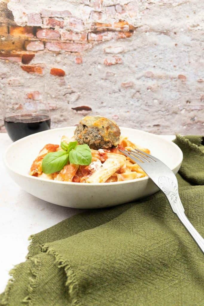 A bowl of delicious vegetable pasta bake, with meatless meatballs, basil and a glass of red wine.