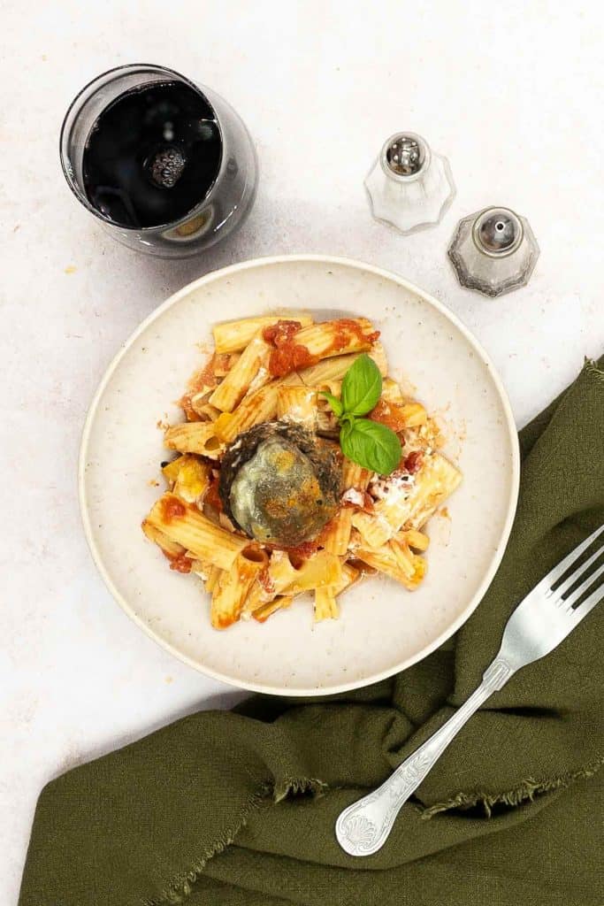 A bowl of vegetarian baked pasta with veggie meatballs,, with rigatoni pasta, a sprig of fresh basil, a fork, salt and pepper and a glass of red wine.