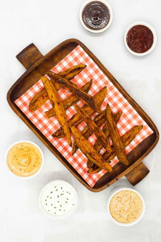 Freshly made roasted sweet potato wedges on a tray with ketchup, barbecue sauce, sour cream and chives, sriracha mayo sauce and chipotle aioli.