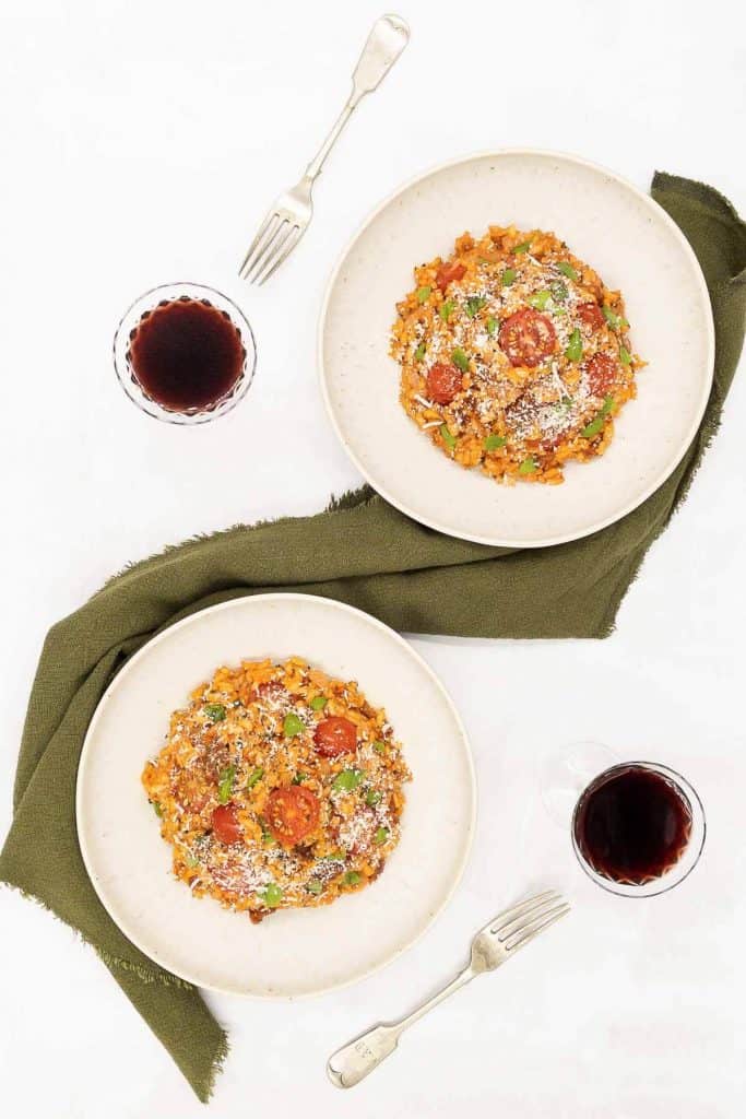Overhead shot of two bowls of tomato vegan risotto with two glasses of red wine and forks.