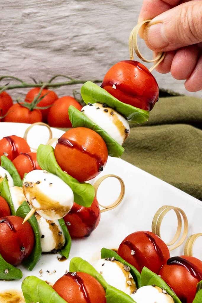 Holiding up a Caprese skewer made from tomato, mozzarella cheese and basil leaves.