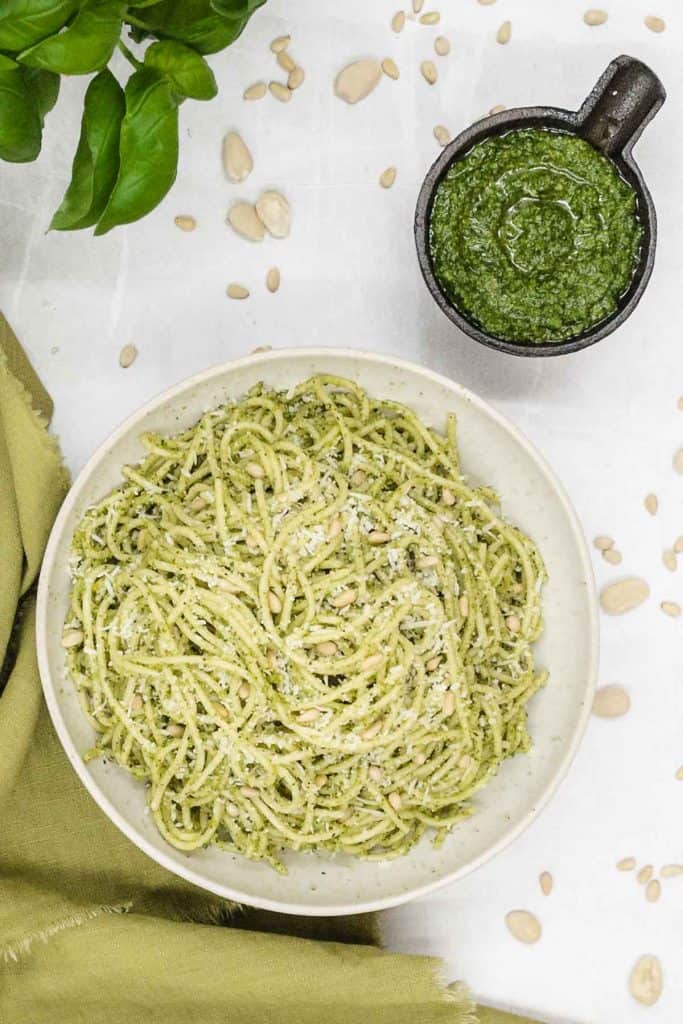 An overhead view of a bowl of warm pasta with a creamy sauce, made from basil pesto, with a mortar of freshly made pesto, fresh herbs and toasted pine nuts.