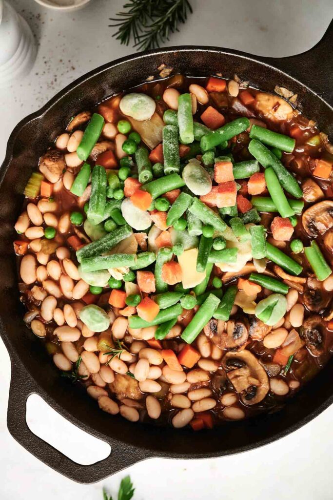 A cast iron pan brimming with a vibrant vegetarian shepherd's pie features mixed vegetables, beans, and mushrooms in a savory sauce, crowned with frozen green beans and carrots.