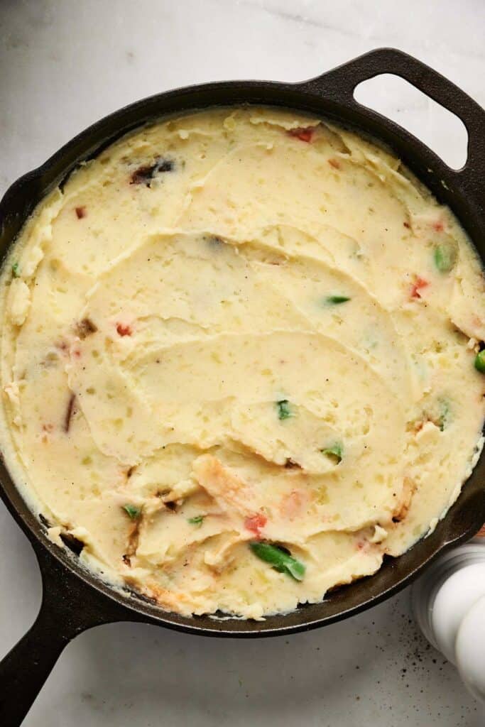 A skillet filled with mashed potatoes mixed with assorted vegetables, including green and red peppers, mimicking a vegetarian shepherd's pie, rests on a marble surface.