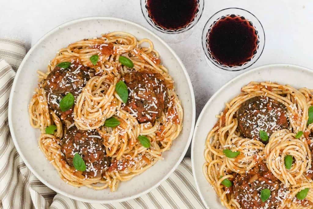 Two bowls of meat free meatballs and spaghetti in a marinara sauce, fresh basil and glasses of red wine.