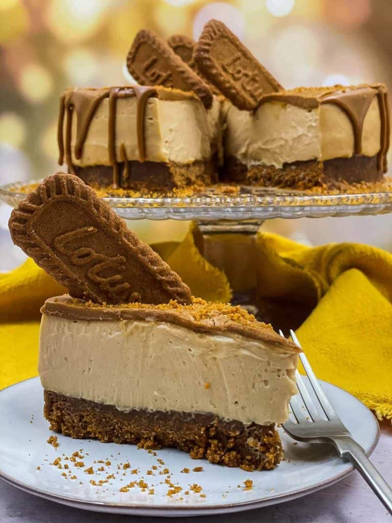 A slice of delicious cheesecake made with Biscoff on a plate with a fork and a cake stand behind it with the rest of the cheesecake on it.