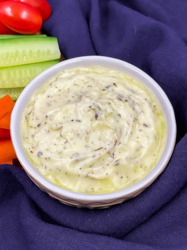 A bowl of garlic mayonnaise herb dip with veggies cut up, cucumbers, carrots and tomatoes.