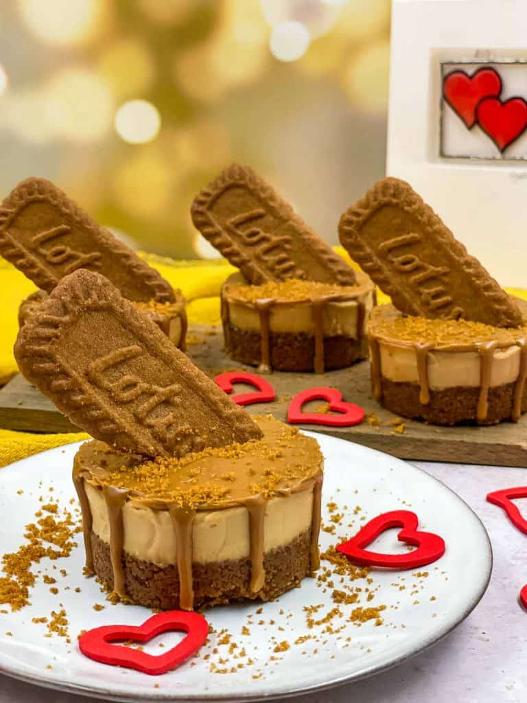 Individual cheesecakes made with Biscoff, one on a plate with crushed biscuits on the plate, the cheesecake has a Biscoff topping which has oozed over the side and there are lotus biscuits stuck into each cheesecake.