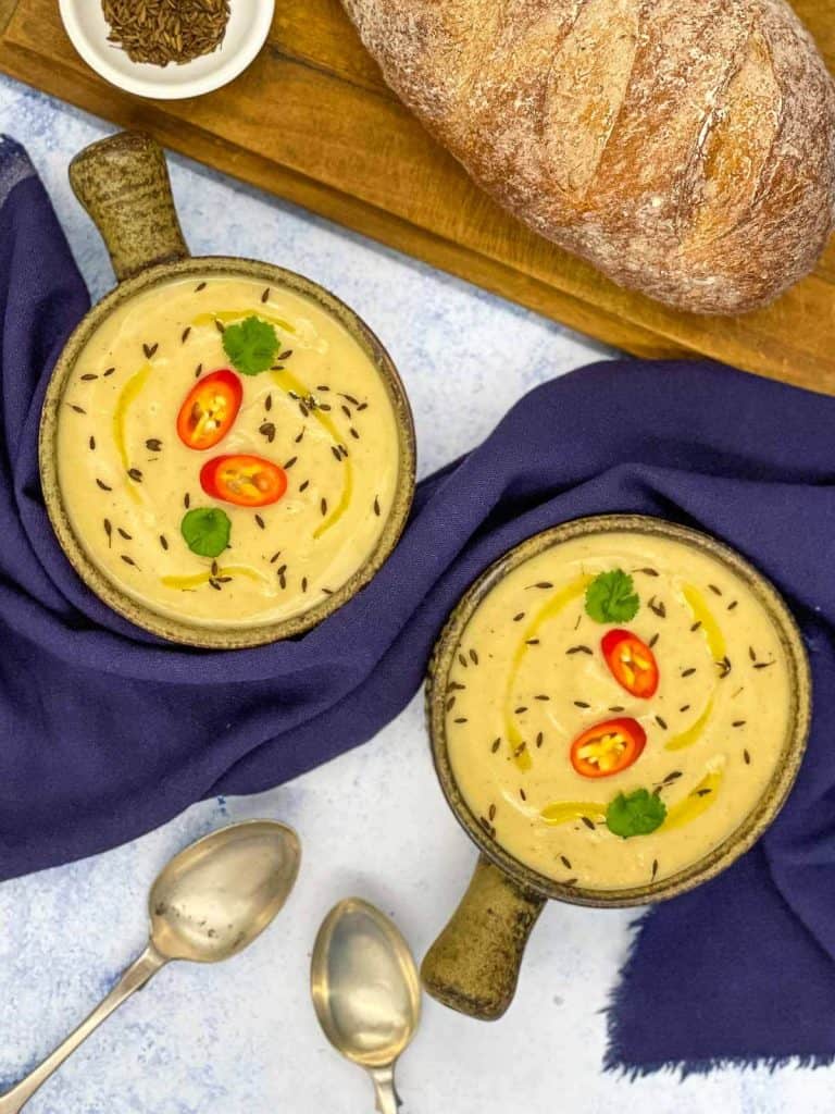 Two spoons underneath two bowls of tasty soup made with parsnips, a loaf of crusty bread and a small bowl of cumin seeds.
