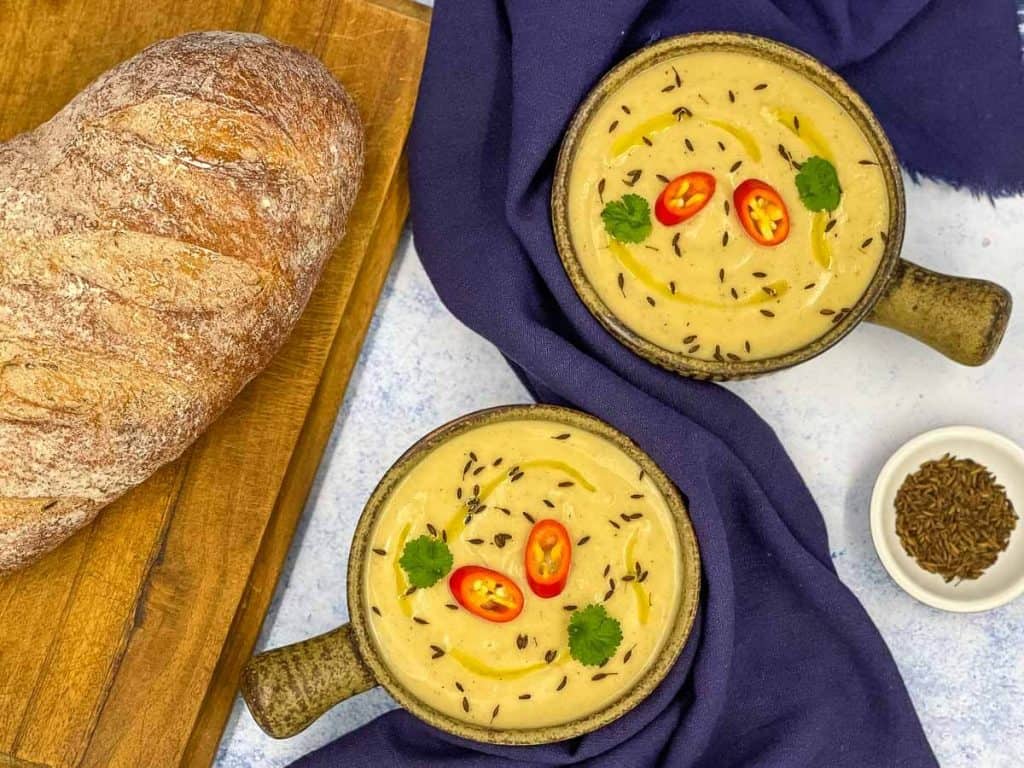 Homemade parsnip soup in bowls with a loaf of crusty bread and a bowl of toasted cumin seeds.