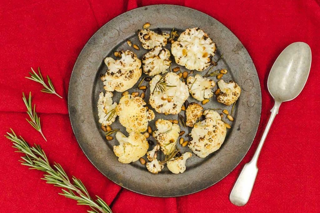 A plate of roasted cauliflower with rosemary, pine nuts and a serving spoon.