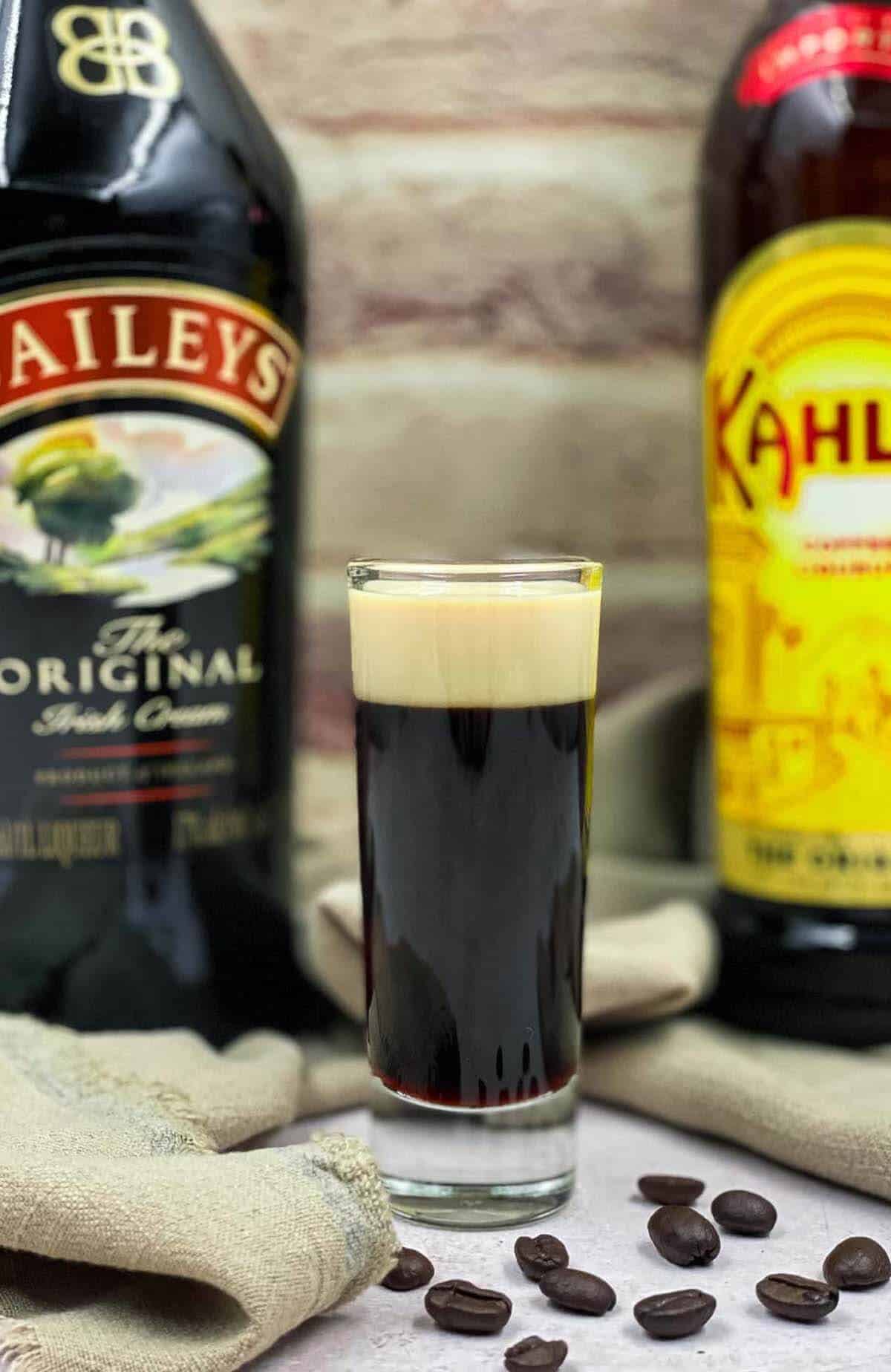 Baby Guinness shot in the middle of two liqueur bottles and coffee beans.