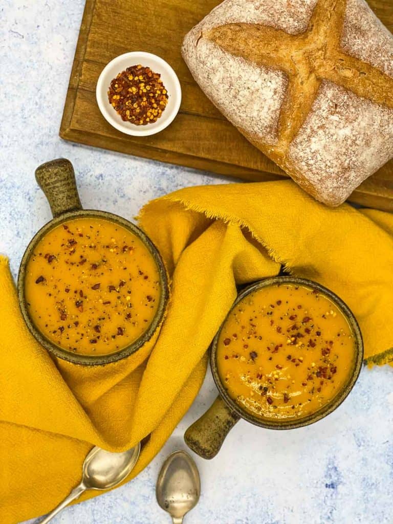 Two bowls of delicious carrot and parsnip soup, with red chili flakes and a loaf of crusty bread.