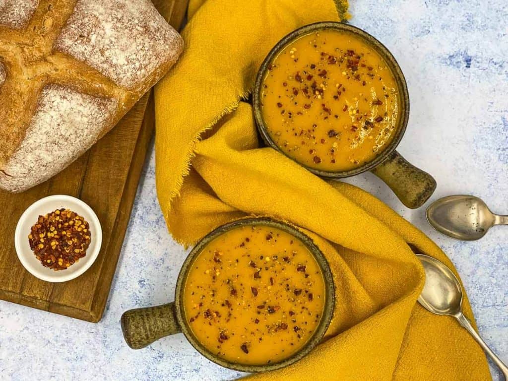 Delicious parsnip and carrot soup, in bowls with red chili flakes and a loaf of crusty bread.