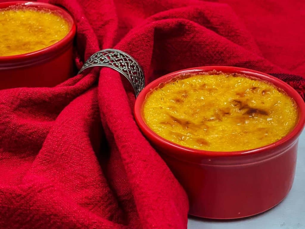 A creme brulee in a red ramekin, with a golden brown caramelized sugar topping, with red table linen and a napkin ring.