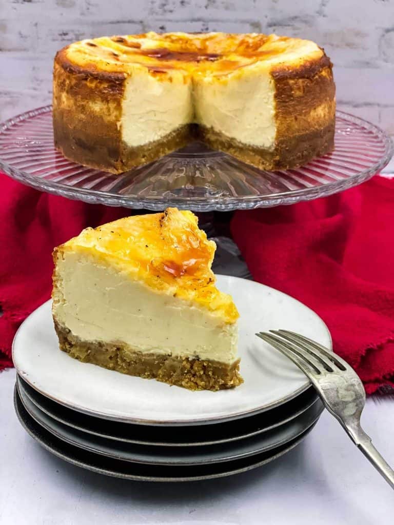 A slice of delicious cheesecake made with in the style of creme brulee on a plate with a fork and a cake stand behind it with the rest of the cheesecake on it.