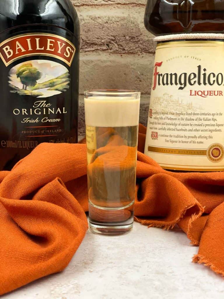 A layered shot of nutty Irishman in a shot glass with Baileys Irish cream and Frangelico liqueur bottles in the background.