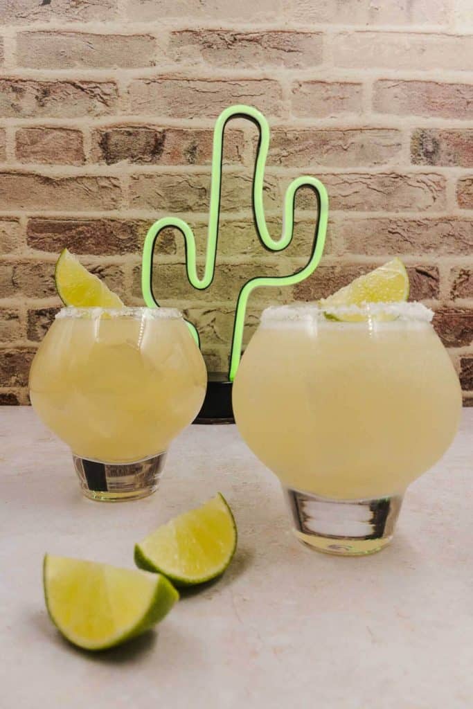 Two glasses of tequila, grapefruit cocktail with lime wedges and a neon cactus in the background.