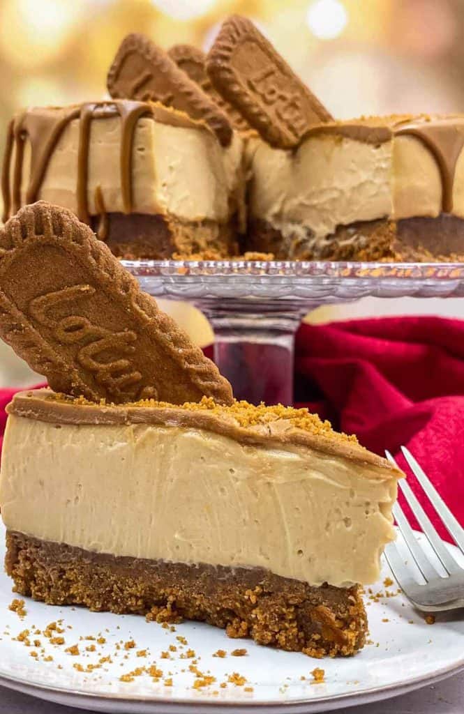 A slice of vegan Biscoff cheesecake on a plate with the rest of the cheesecake on a cake stand.
