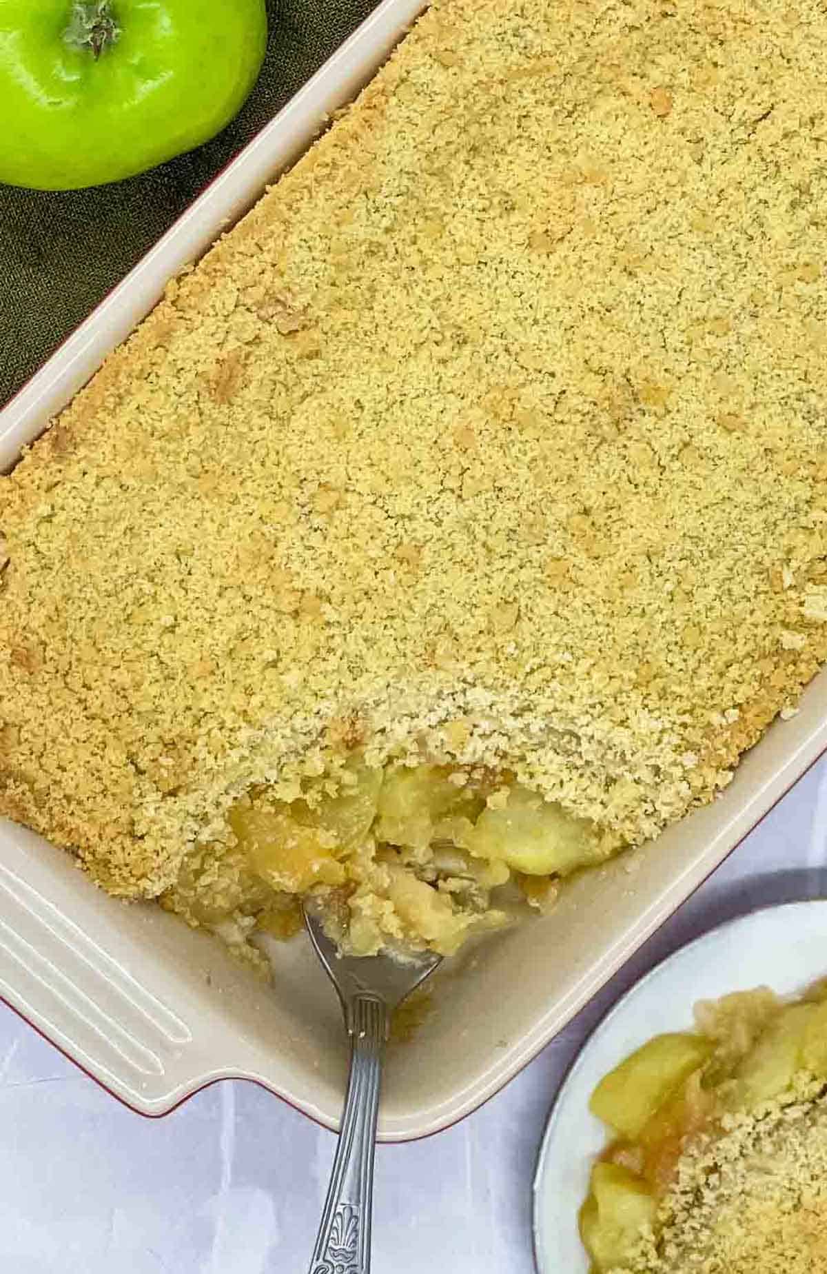 Apple crumble in a baking dish with a portion on a plate and serving spoon in the baking dish.