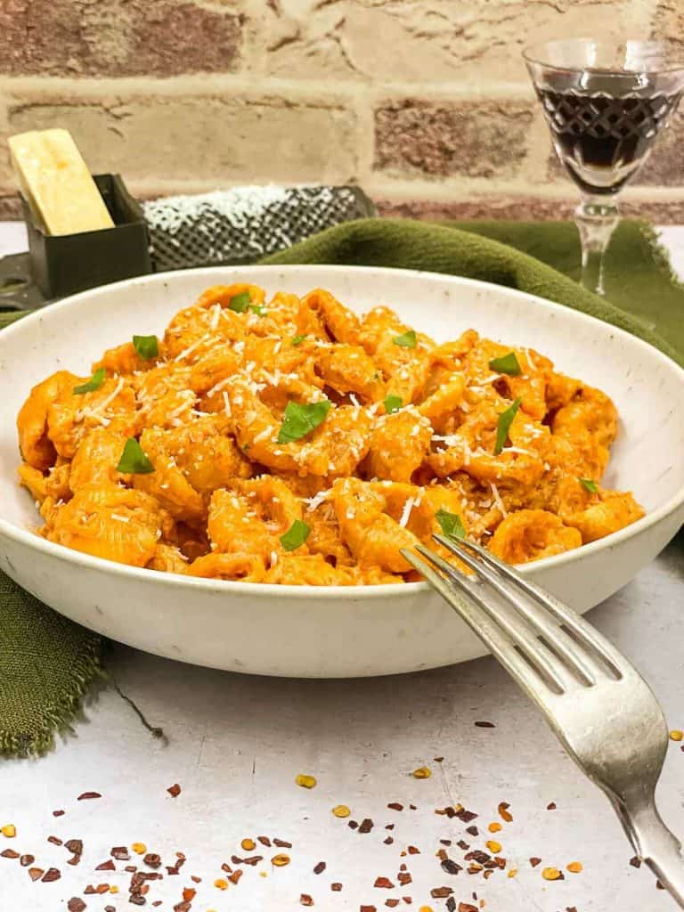 A bowl of creamy, spicy, vodka pasta with a fork resting on the side, a glass of red wine, red pepper flakes, and Parmesan cheese in a grater.