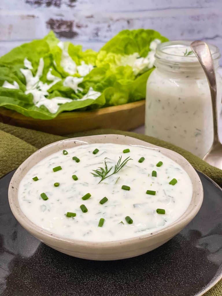 A bowl of homemade ranch dressing with chives and dill garnish, a jar of dressing in the background and used as salad dressing on lettuce.