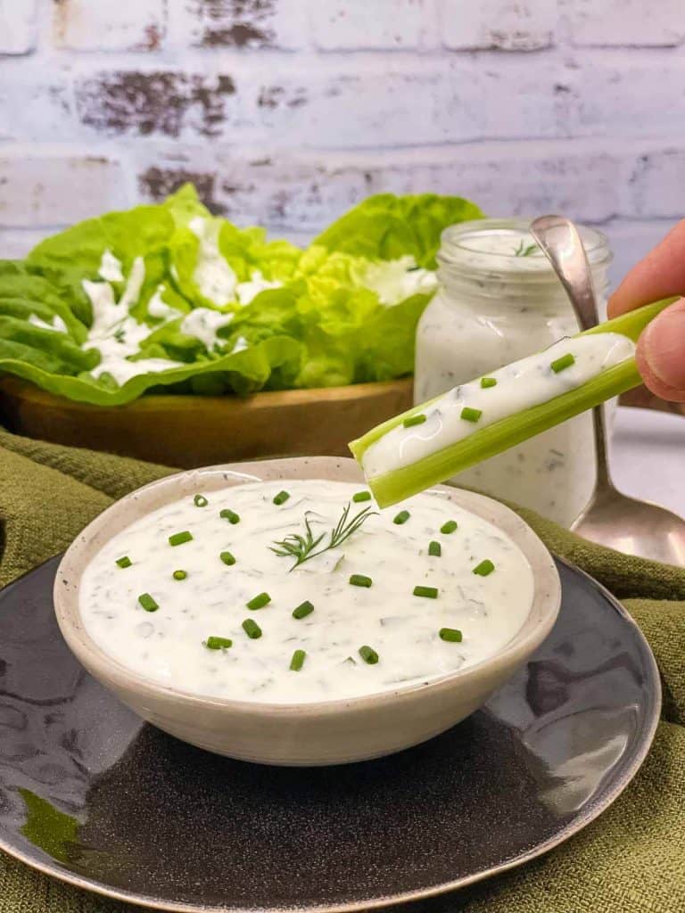 A person dipping into the ranch dressing with a celery stick, a jar of salad dressing in the background and lettuce with it drizzled on.
