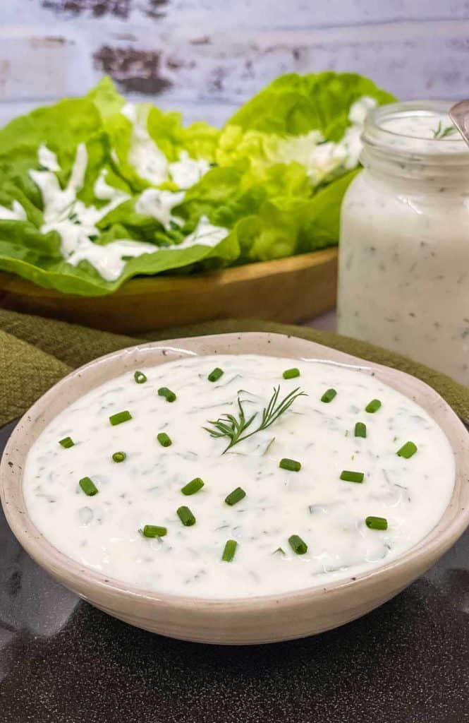A bowl of fresh ranch dressing with chives and dill dressing, a jar full of dressing and lettuce leaves with ranch on them in the background.