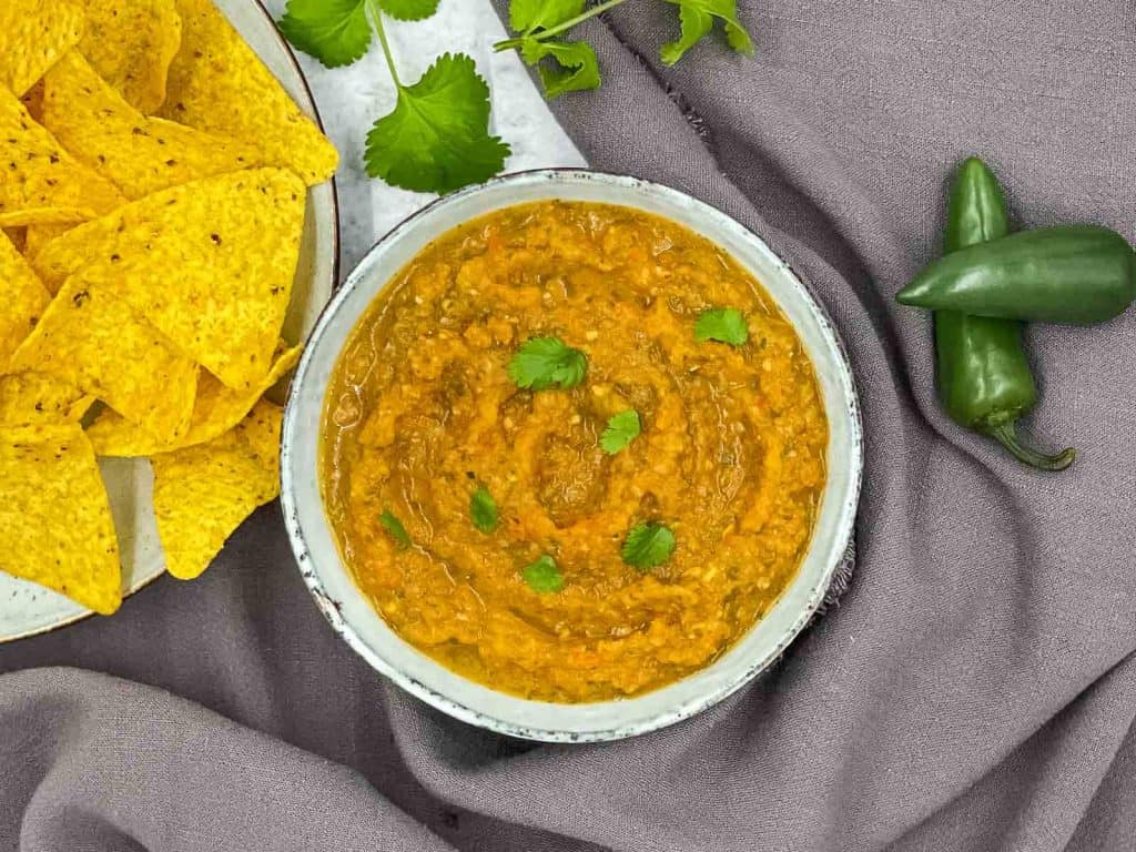 A bowl of salsa Roja, with tortilla chips, cilantro and jalapeno peppers.