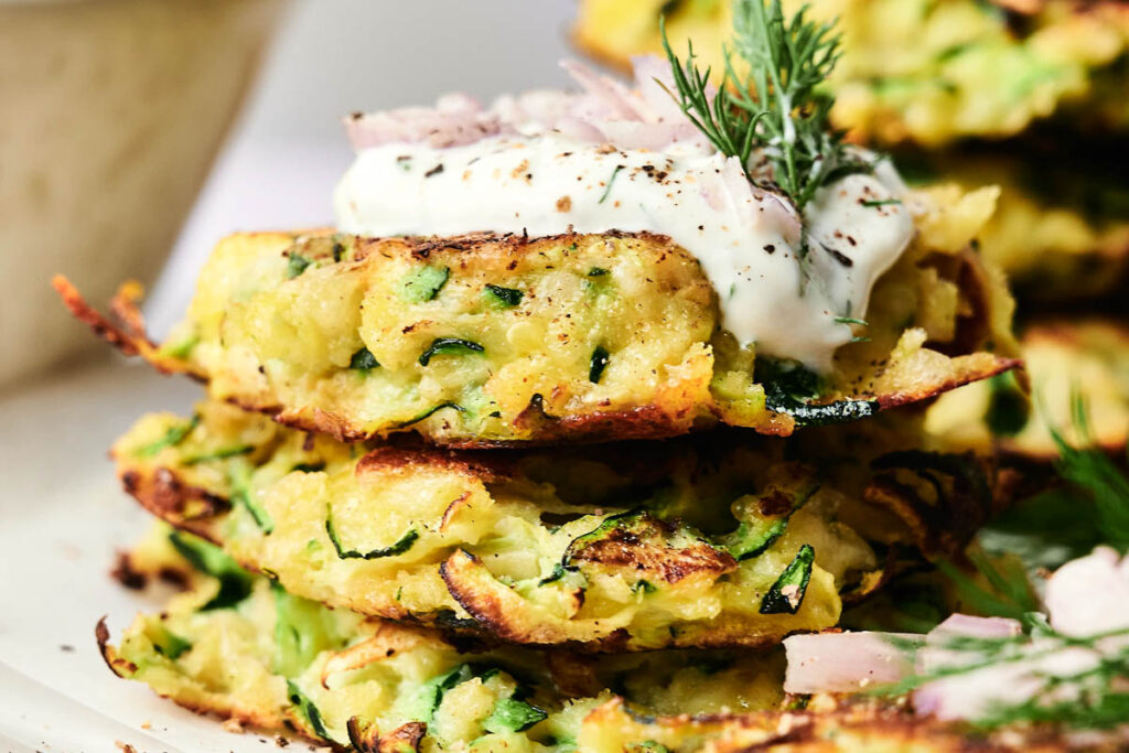 A stack of crispy zucchini fritters topped with a dollop of sour cream, chopped onions, and fresh dill.