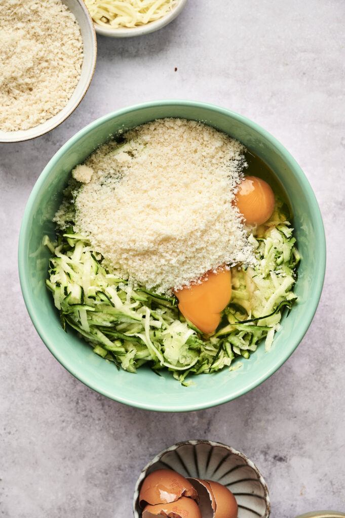 A bowl with grated zucchini, eggs, and breadcrumbs on a light surface—perfect for making crisp zucchini fritters—with additional bowls of breadcrumbs and eggshells nearby.