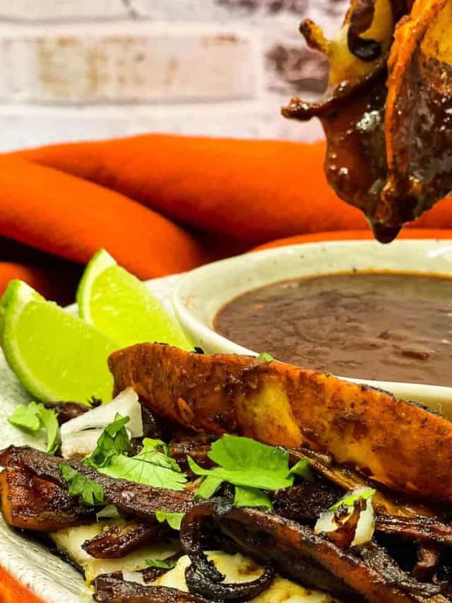 A close-up of a plate with vegan quesabirria tacos filled with seasoned mushrooms, lime wedges, chopped onions, cilantro, and a bowl of consomm&eacute; for dipping.