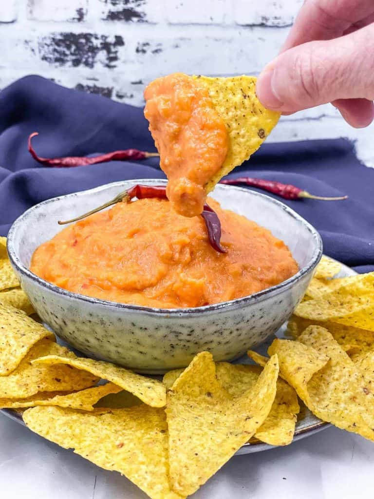 A bowl of delicious salsa tequera with someone dipping in a tortilla chip, with torilla chips around the plate, and Arbol chili peppers.