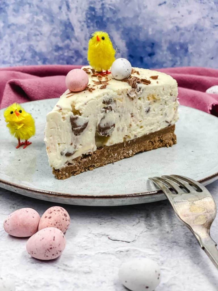 A slice of cheesecake with mini eggs and a chick toy standing on top.