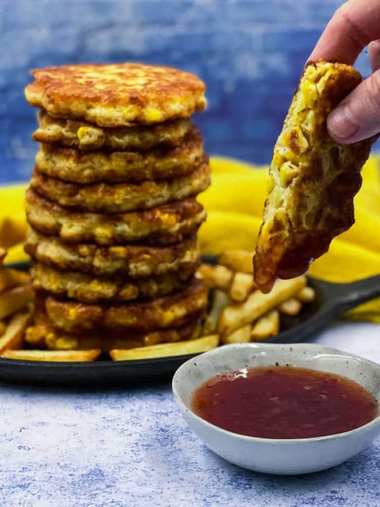 Someone dipping half a corn fritter in sauce with more sweetcorn fritters in the background.