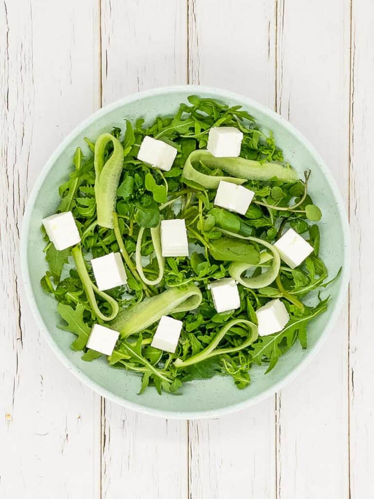 Rocket, watercress, cucumber ribbons, and feta cheese in a bowl.