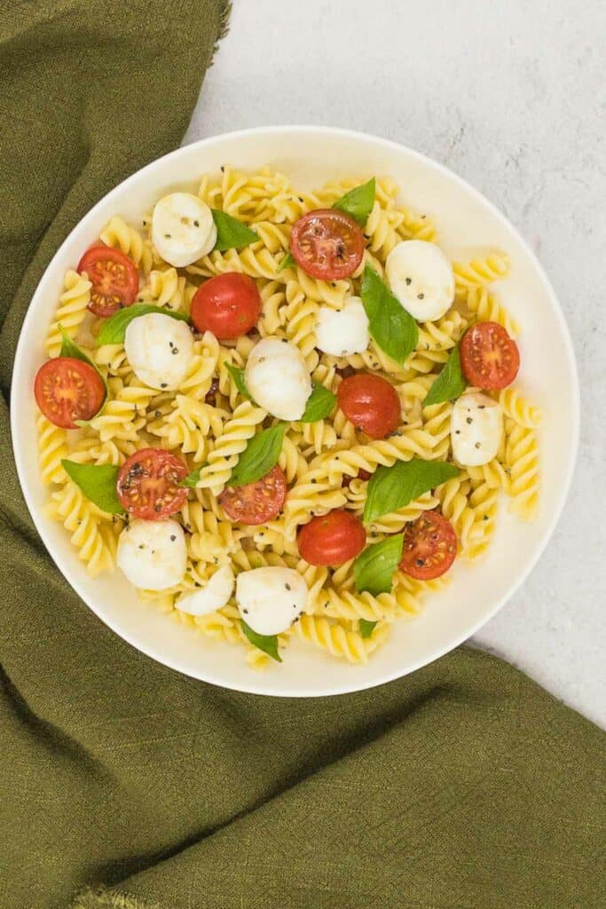 Pasta, mozzarella pearls, cherry tomatoes, fresh basil leaves, salt, and pepper in a bowl.